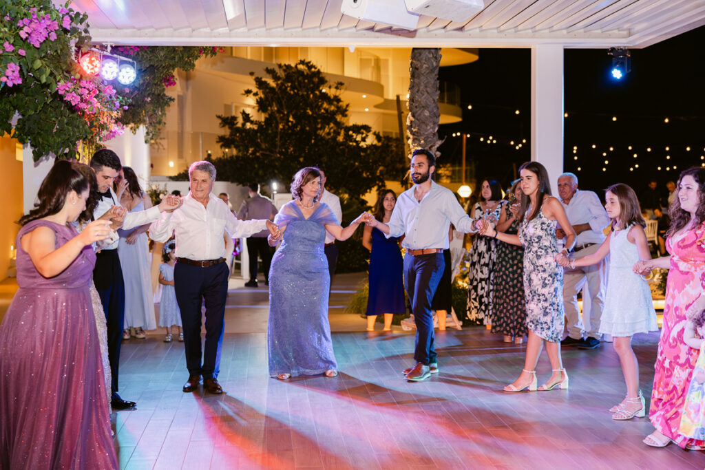 People dressed formally holding hands and dancing in a circle on an outdoor venue with lights.