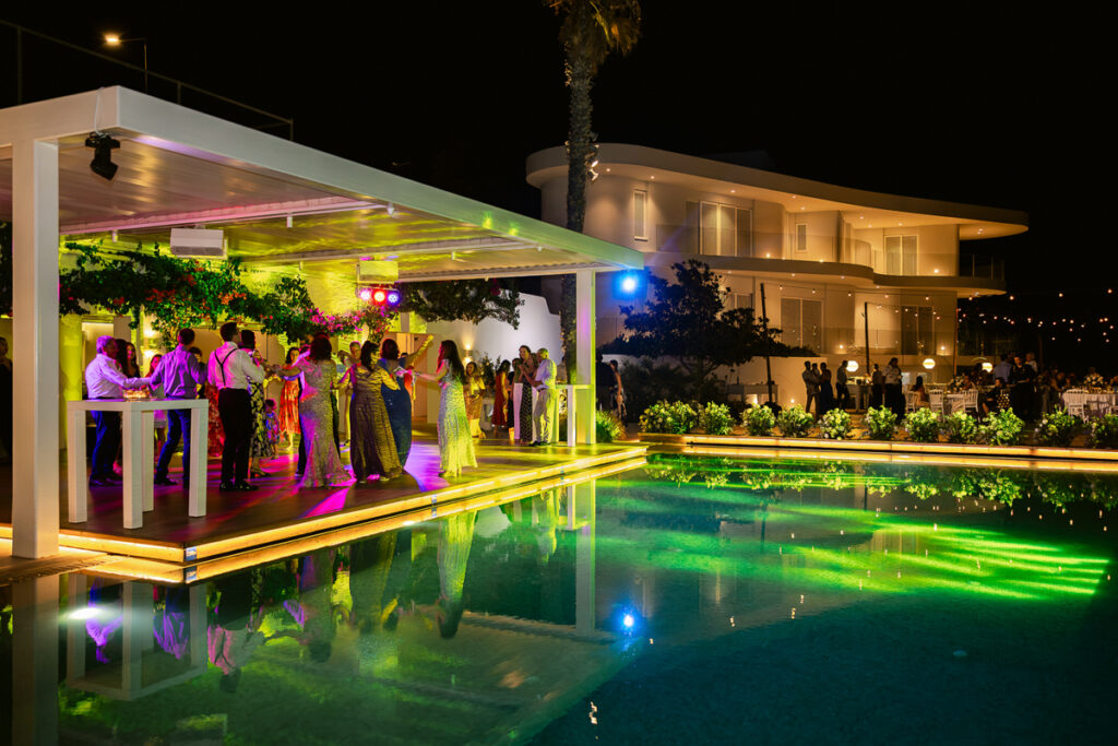 Nighttime party by a pool with people dancing under a lit patio and guests near tables.