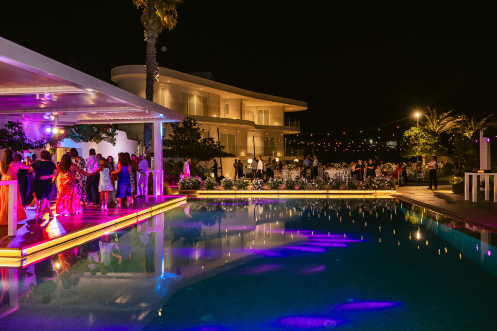 Nighttime outdoor event by a pool with purple lighting, guests on deck and seated at tables.
