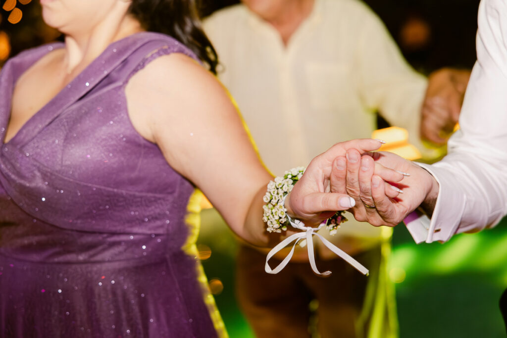 Two people holding hands, one wearing a purple dress and floral wrist corsage, the other in white shirt with wedding ring.