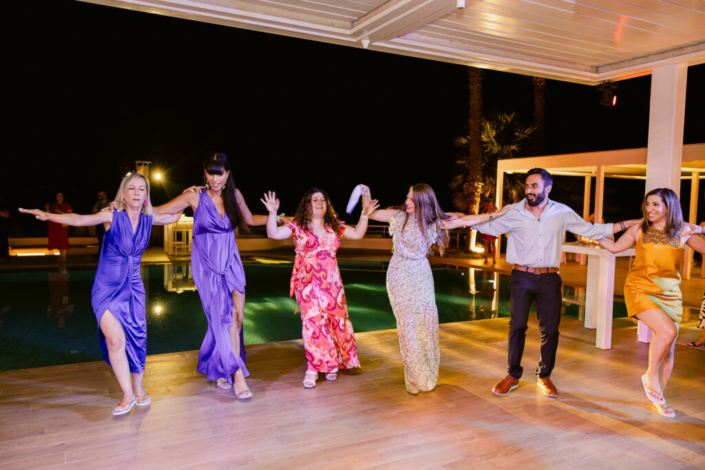 Six people holding hands and dancing outdoors at night near a pool on a patio.