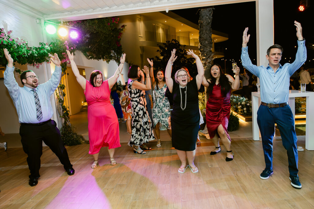 Seven people in formal and semi-formal clothes dancing with raised arms on wooden floor at night.
