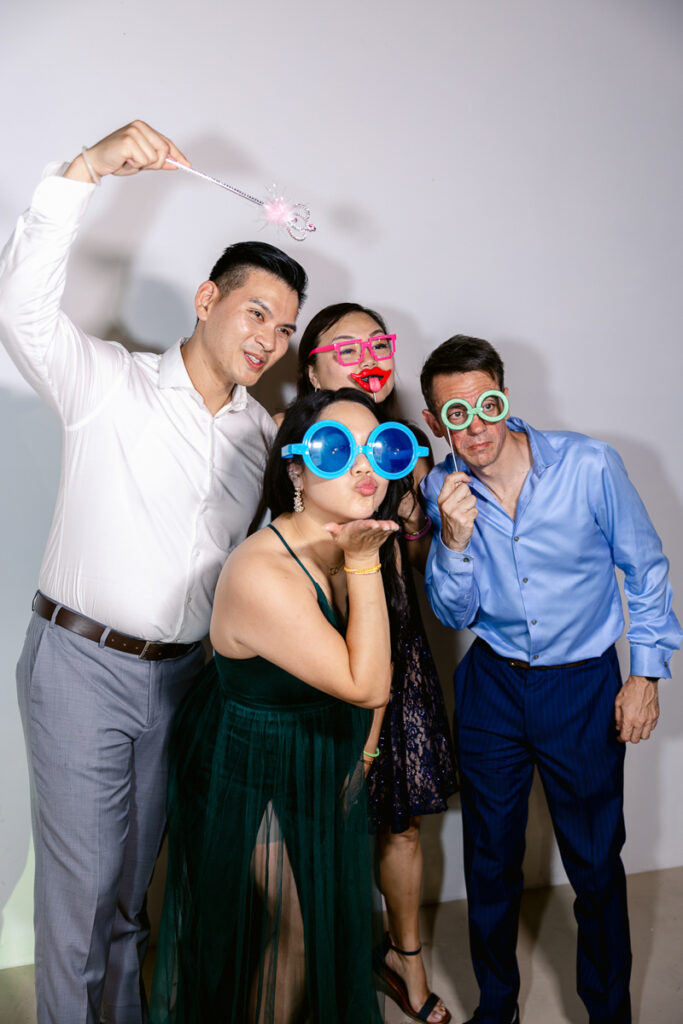 Four adults wearing colorful oversized glasses and holding props pose against a plain wall.