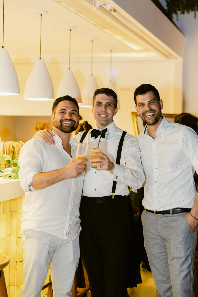 Three men indoors holding drinks, one in formal attire with suspenders and bow tie.