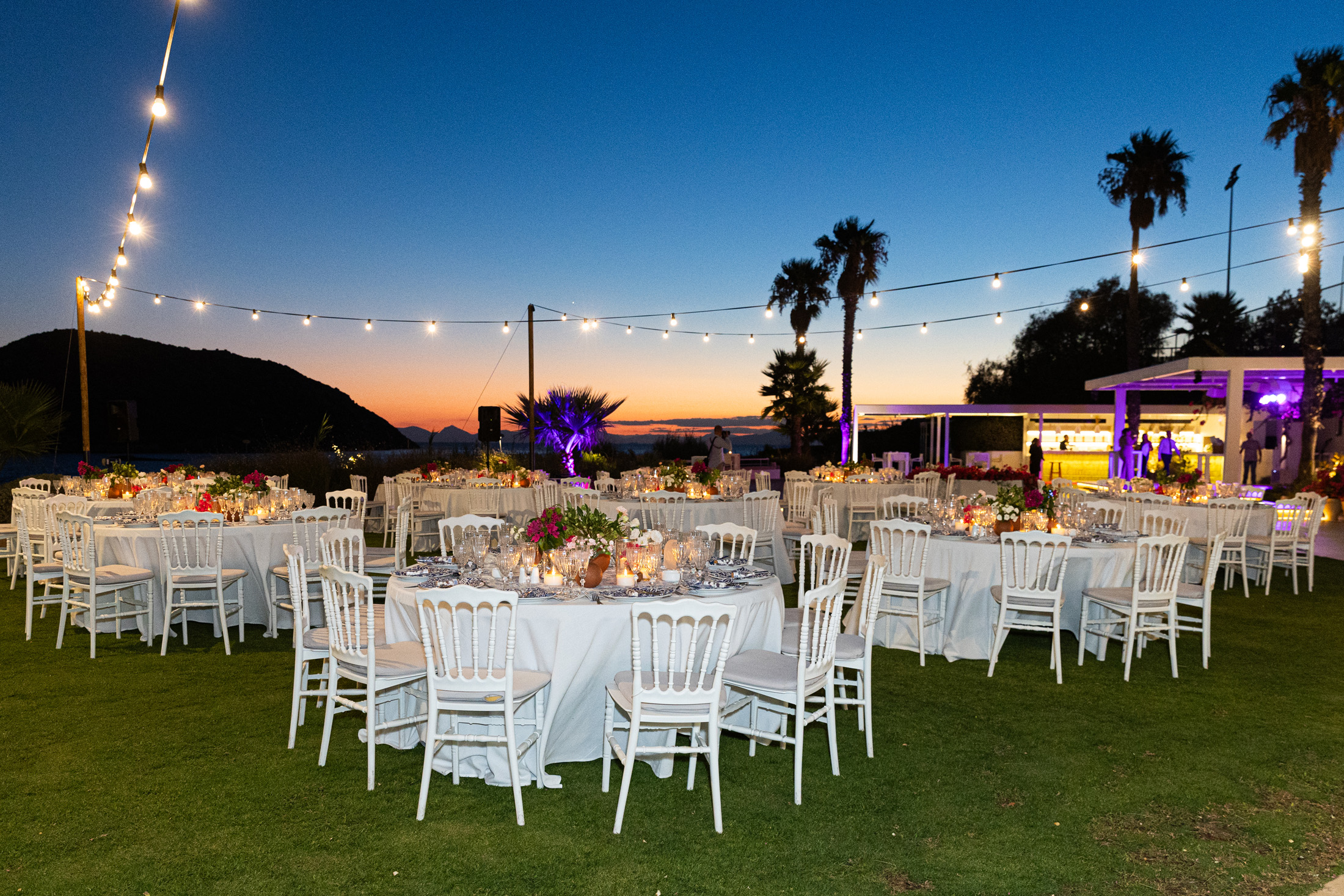 Outdoor event setup with round tables, white chairs, string lights, and sunset sky.